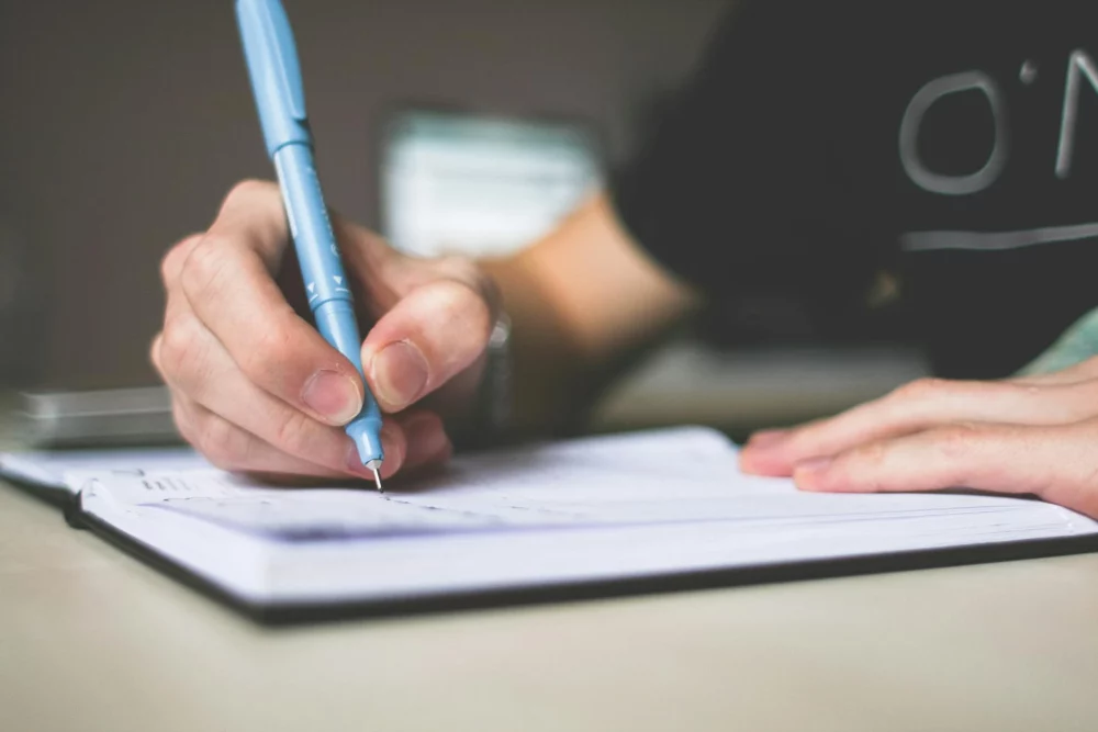 A close-up shot of a person writing in a notebook with a blue pen expressing their grief.